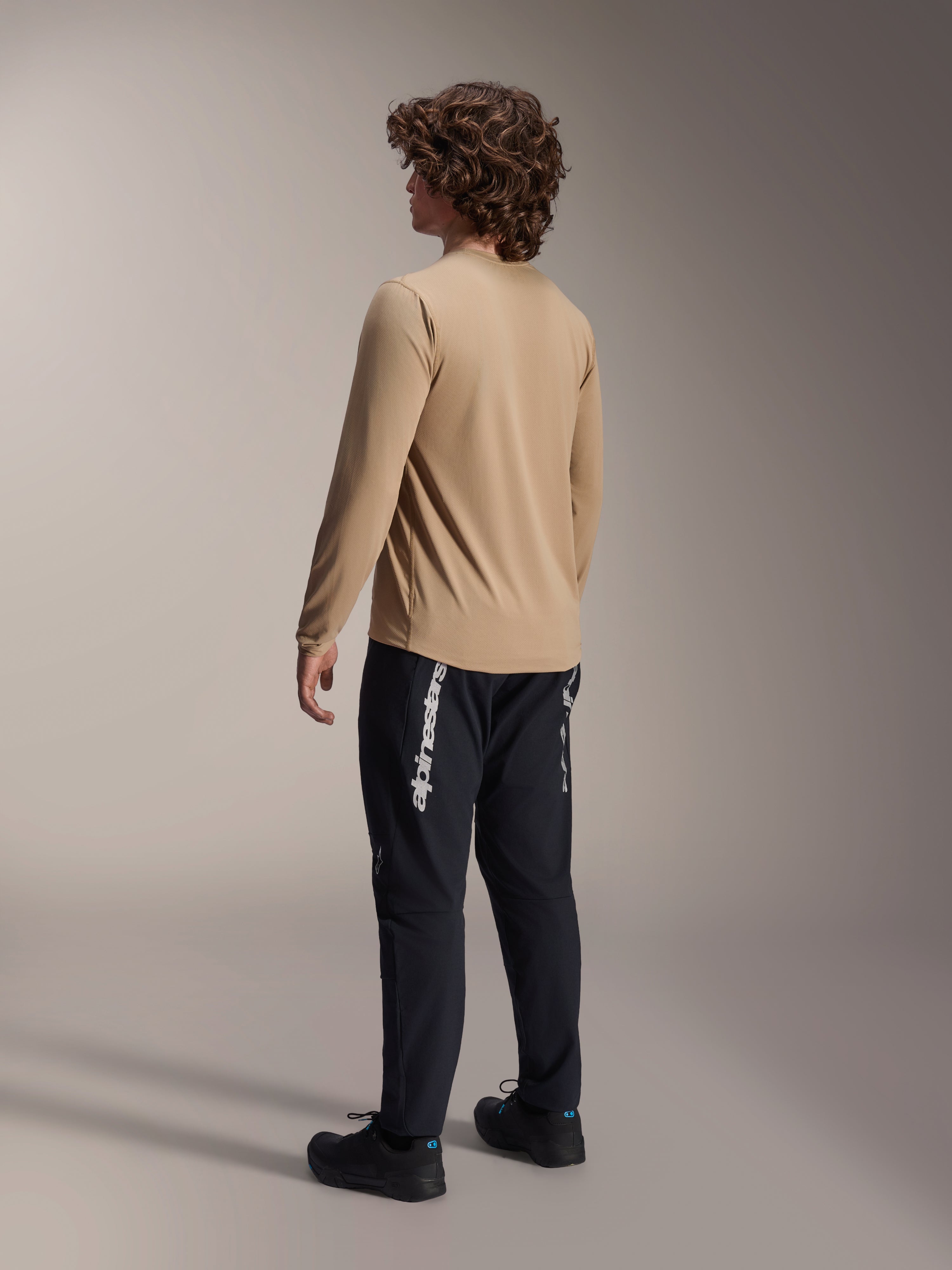 Maglia a maniche lunghe Alpinestars A-Dura Astar in marrone chiaro, vista posteriore di un modello maschile che mostra il design a coda allungata, abbinata a pantaloni e scarpe neri, abbigliamento tecnico casual per mountain bike.