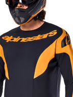 Alpinestars A-Supra Race Jersey - Manica Lunga, nera con inserti grafici arancioni, grande marchio Alpinestars arancione sul petto, indossata da un pilota con casco integrale coordinato nero e arancione, abbigliamento tecnico da gara MTB