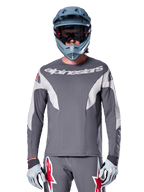 Ciclista MTB che indossa la maglia Alpinestars A-Supra Race - Manica Lunga in grigio scuro, maglia da ciclismo a maniche lunghe con logo Alpinestars bianco, abbinata a casco integrale grigio-azzurro, maschera e guanti da corsa grigi, equipaggiamento completo da gara per mountain bike per prestazioni off-road