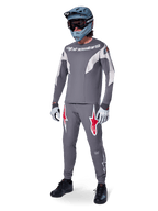 Pilota di MTB con maglia Alpinestars A-Supra Race a maniche lunghe, grigio scuro con dettagli grigio chiaro, pantaloni coordinati con logo a stella rosso, casco integrale grigio con maschera e guanti, completo tecnico per gare di mountain bike
