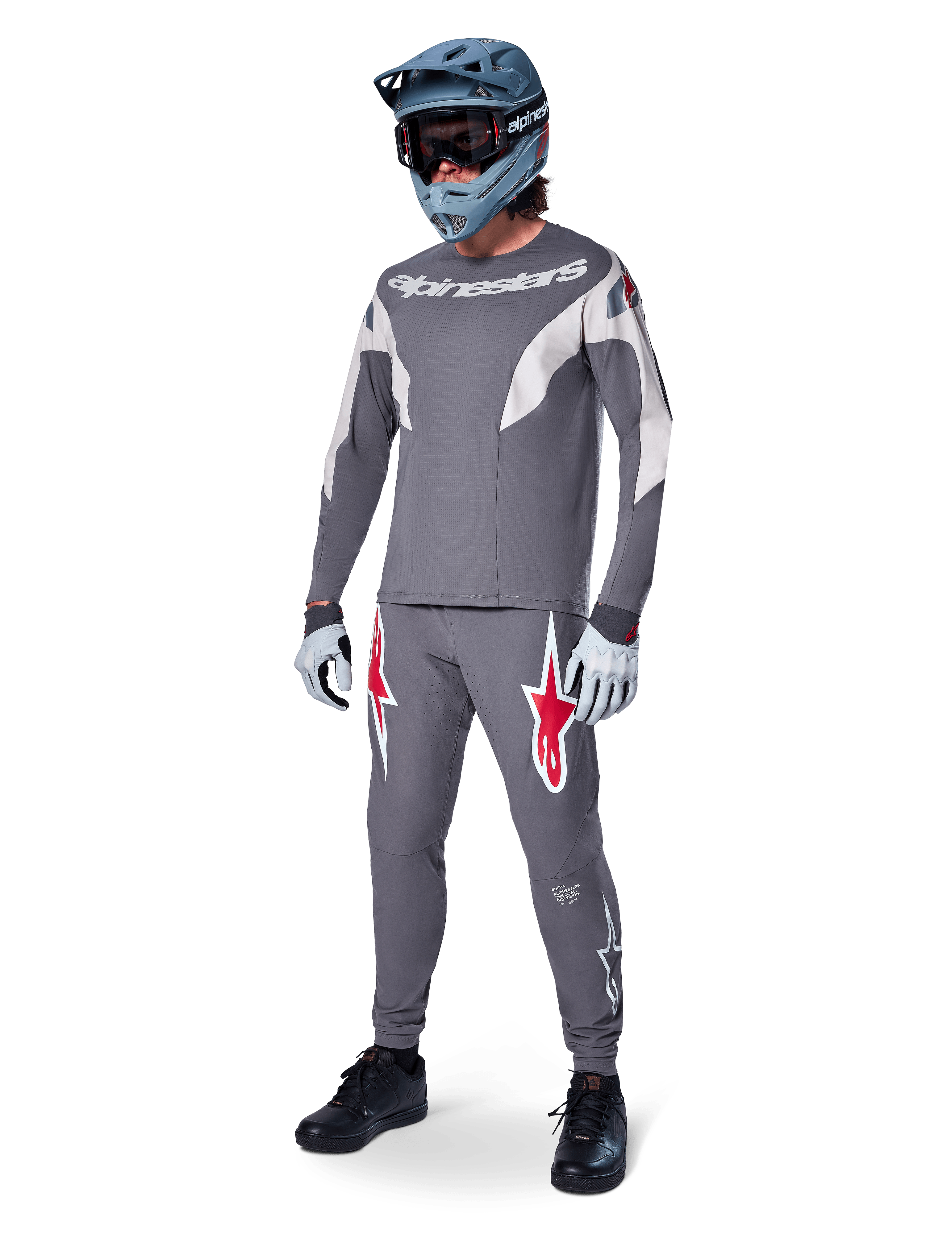 Pilota di MTB con maglia Alpinestars A-Supra Race a maniche lunghe, grigio scuro con dettagli grigio chiaro, pantaloni coordinati con logo a stella rosso, casco integrale grigio con maschera e guanti, completo tecnico per gare di mountain bike
