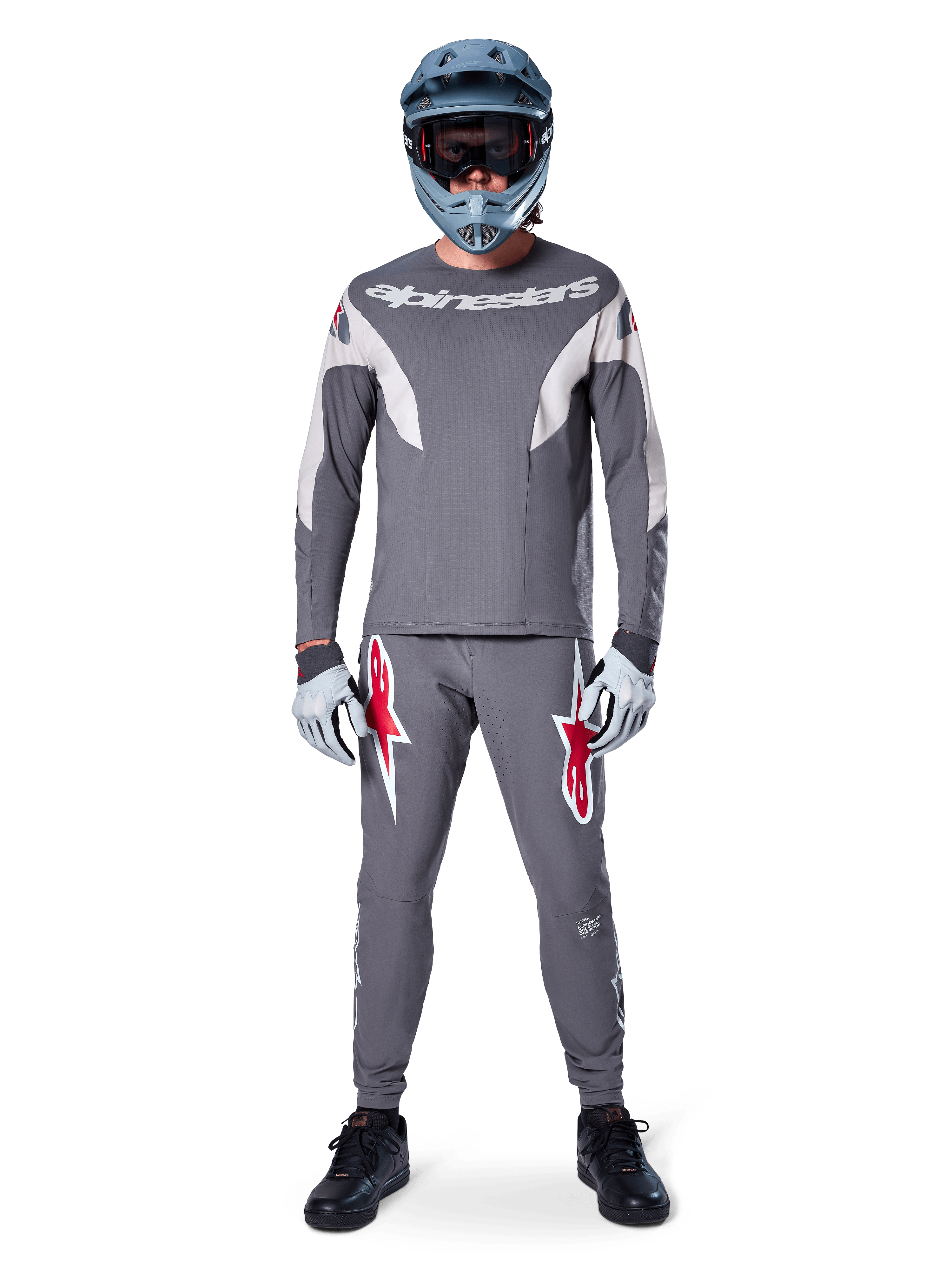 Rider con maglia Alpinestars A-Supra Race - Manica lunga in grigio scuro, coordinata con pantaloni grigi abbinati con vistosi loghi a stella rossi, casco grigio con maschera e guanti grigio chiaro, kit da gara MTB completo per il ciclismo fuoristrada ad alte prestazioni