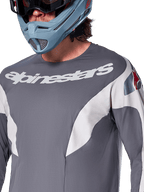 Alpinestars A-Supra Race Jersey - a maniche lunghe, maglia MTB, grigio scuro con grafiche racing bianche e logo Alpinestars bianco, indossata con casco integrale azzurro e maschera, tessuto microperforato per prestazioni off-road