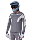 Alpinestars A-Supra Race Jersey a maniche lunghe, grigio scuro con dettagli bianchi e rossi, grafiche in stile racing, indossata con pantaloni grigi coordinati, casco integrale blu e guanti bianchi, equipaggiamento professionale per MTB e downhill