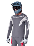 Alpinestars A-Supra Race Jersey a maniche lunghe, grigio scuro con dettagli bianchi e rossi, grafiche in stile racing, indossata con pantaloni grigi coordinati, casco integrale blu e guanti bianchi, equipaggiamento professionale per MTB e downhill