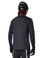 Maglia a maniche lunghe Alpinestars A-Dura Switch, nera, vista posteriore su un ciclista con casco MTB nero e guanti, in tessuto tecnico jacquard e con coda allungata per il fuoristrada