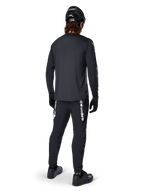 Maglia a maniche lunghe Alpinestars A-Dura Switch, nero, vista posteriore su un ciclista, indossata con pantaloni neri con logo Alpinestars bianco, casco nero e guanti, dotata di coda allungata per maggiore copertura, abbigliamento tecnico da mountain bike