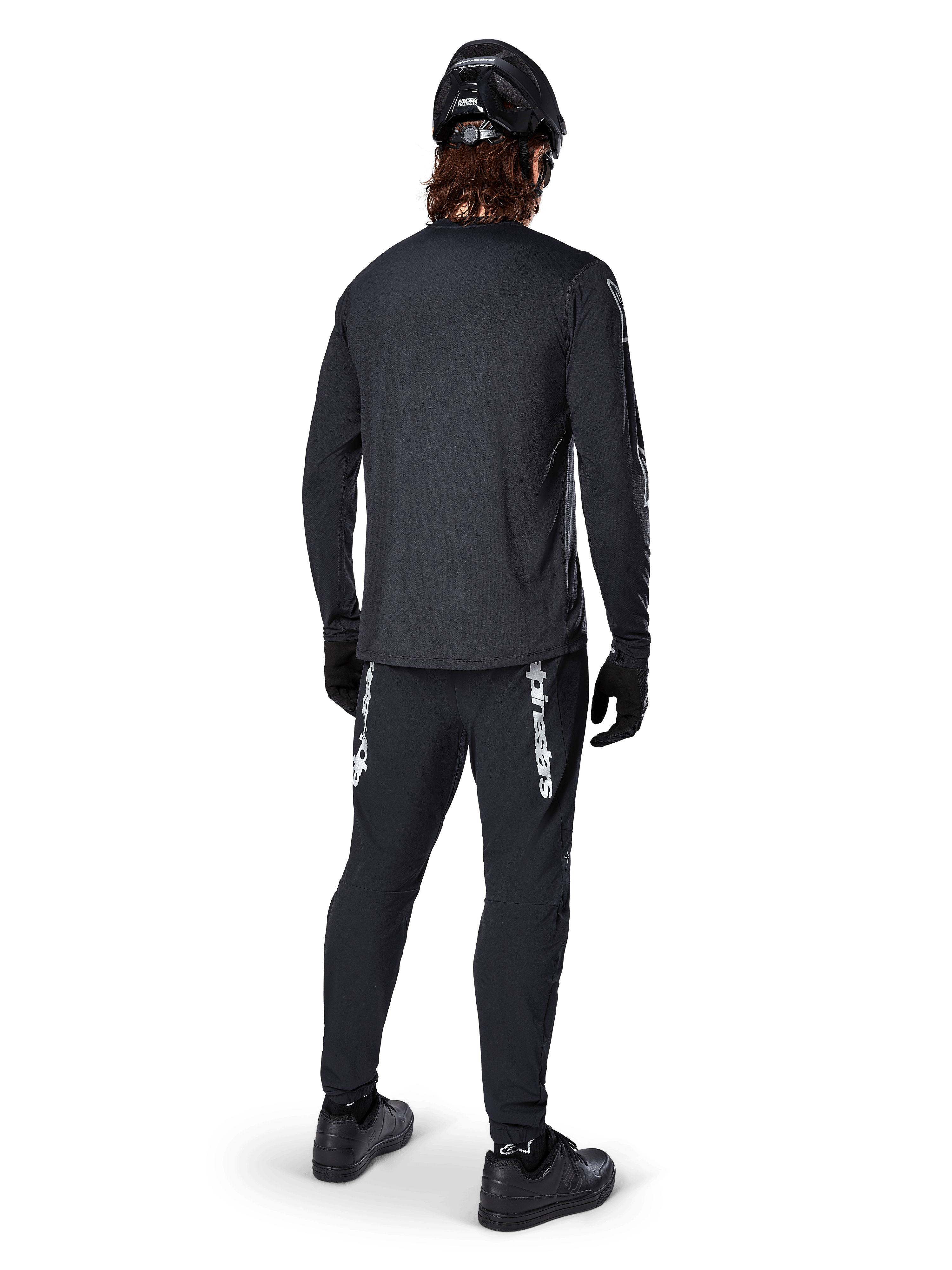 Maglia a maniche lunghe Alpinestars A-Dura Switch, nero, vista posteriore su un ciclista, indossata con pantaloni neri con logo Alpinestars bianco, casco nero e guanti, dotata di coda allungata per maggiore copertura, abbigliamento tecnico da mountain bike