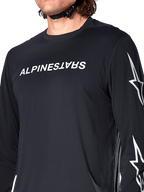 Maglia Alpinestars A-Dura Switch Long Sleeve, nero, con scritta Alpinestars bianca sul petto e loghi a stella sulla manica, indossata da un rider con cinturino del casco visibile, abbigliamento tecnico da mountain bike.