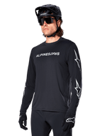 Ciclista uomo che indossa la maglia a maniche lunghe Alpinestars A-Dura Switch, nero con scritta bianca e loghi a stella sulle maniche, abbinata a casco MTB nero e occhiali da sole, abbigliamento da ciclismo ad alte prestazioni per la guida fuoristrada