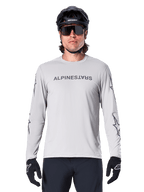 Maglia Alpinestars A-Dura Switch a maniche lunghe, grigio chiaro, con scritta sul petto e loghi a stella sulle maniche in grigio scuro, indossata da un ciclista con casco da MTB e guanti neri, abbigliamento tecnico da ciclismo per trail