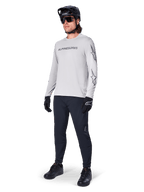 Maglia a maniche lunghe Alpinestars A-Dura Switch, Grigio Chiaro, indossata da un ciclista con pantaloni da mountain bike neri e casco, con logo wordmark nero sul petto e grafiche a stella sulle maniche, abbigliamento tecnico per ciclismo fuoristrada