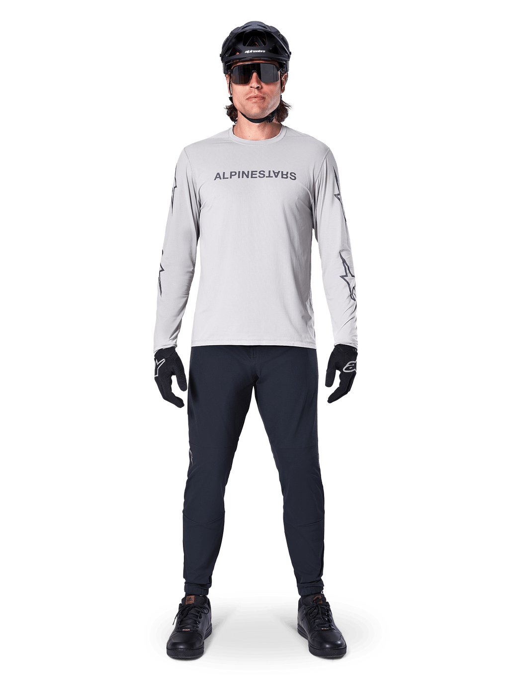 Maglia a maniche lunghe Alpinestars A-Dura Switch in grigio chiaro, con logo wordmark stilizzato sul petto, indossata con pantaloni neri, casco e guanti, abbigliamento tecnico da mountain bike per ciclismo off-road