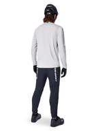 Maglia a maniche lunghe Alpinestars A-Dura Switch, Grigio Chiaro, vista posteriore su un ciclista che indossa pantaloni da ciclismo neri, casco e guanti, abbigliamento da mountain bike ad alte prestazioni con coda posteriore più lunga.