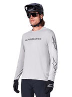 Alpinestars A-Dura Switch Long Sleeve Jersey, maglia da mountain bike, grigio chiaro, logo testuale nero serigrafato sul petto e loghi a stella sulle maniche, indossata con casco da mountain bike nero, occhiali da sole scuri e guanti neri, abbigliamento sportivo per ciclismo fuoristrada