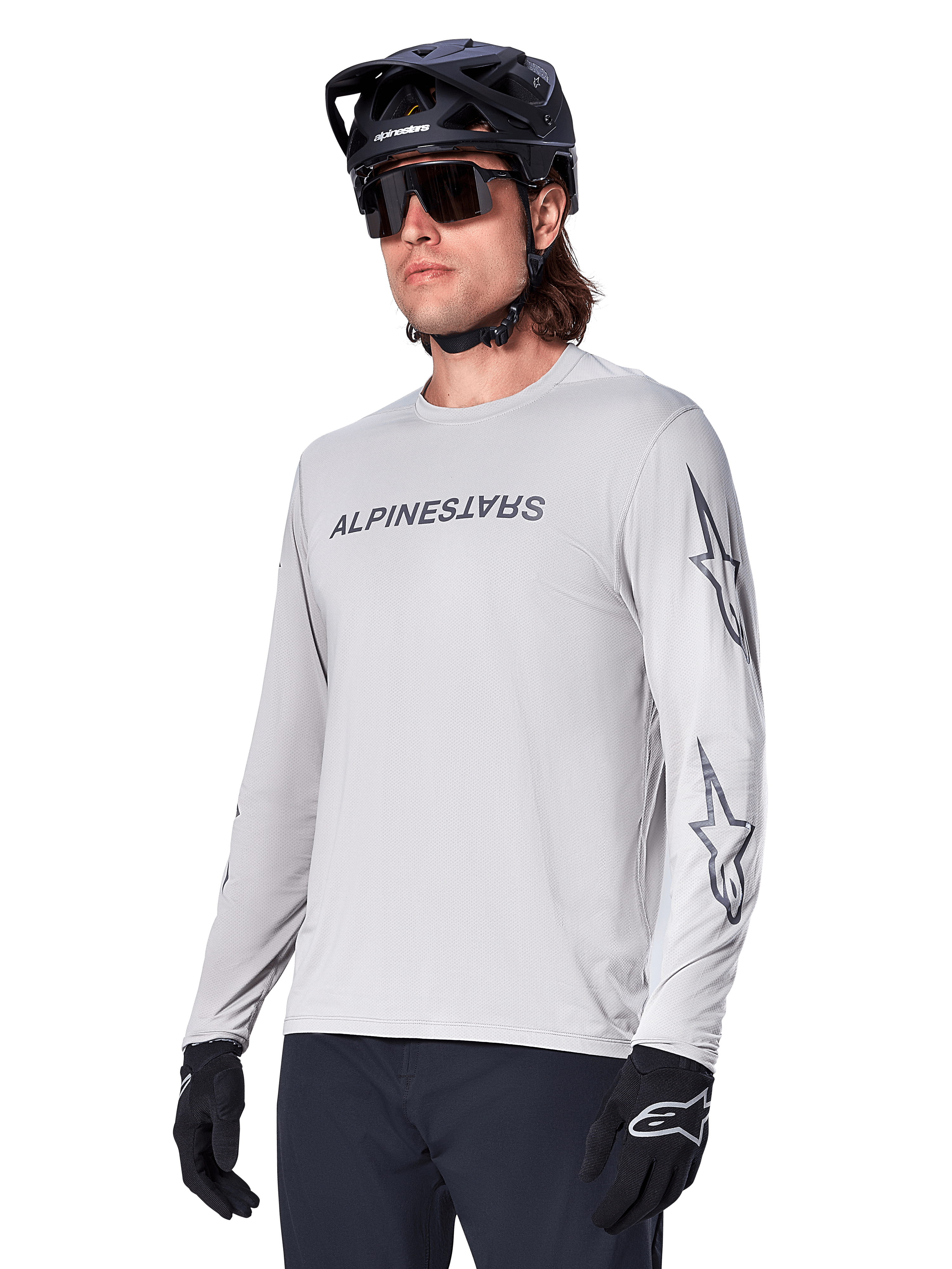 Alpinestars A-Dura Switch Long Sleeve Jersey, maglia da mountain bike, grigio chiaro, logo testuale nero serigrafato sul petto e loghi a stella sulle maniche, indossata con casco da mountain bike nero, occhiali da sole scuri e guanti neri, abbigliamento sportivo per ciclismo fuoristrada