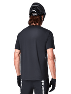 Alpinestars A-Dura Switch Short Sleeve Jersey, nero, vista posteriore su un modello con casco da ciclismo nero e pantaloncini da MTB, mette in risalto la vestibilità regolare e la coda più lunga specifica per la mountain bike