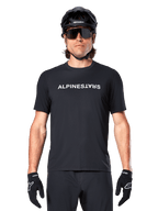 Ciclista di mountain bike con maglia a maniche corte Alpinestars A-Dura Switch in nero, con logo wordmark bianco sul petto, abbinata a casco nero, grandi occhiali da sole e guanti neri, abbigliamento tecnico da ciclismo per trail riding.