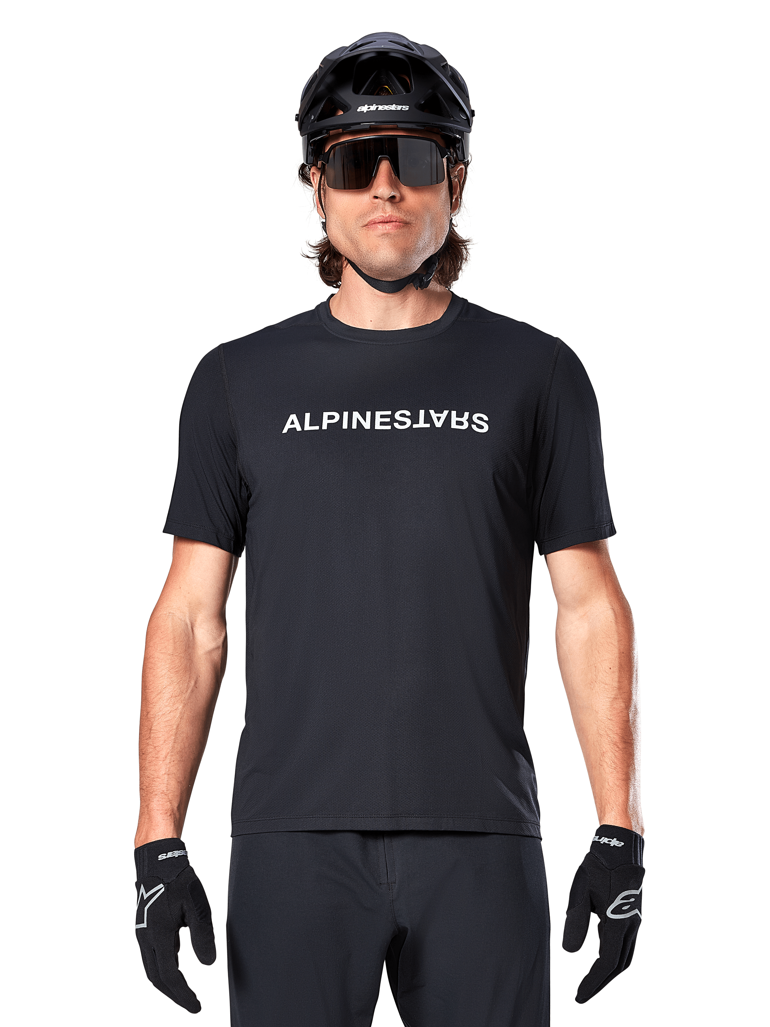 Ciclista di mountain bike con maglia a maniche corte Alpinestars A-Dura Switch in nero, con logo wordmark bianco sul petto, abbinata a casco nero, grandi occhiali da sole e guanti neri, abbigliamento tecnico da ciclismo per trail riding.