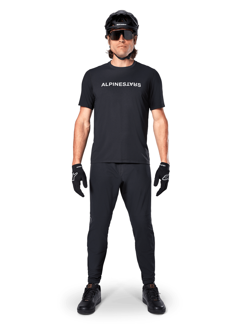 Ciclista di mountain bike che indossa la maglia Alpinestars A-Dura Switch a maniche corte in nero, con logo a scritta bianco sul petto, abbinata a pantaloni da MTB, casco e guanti neri coordinati, completo da ciclismo pronto per il trail.