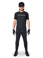 Ciclista di mountain bike che indossa la maglia Alpinestars A-Dura Switch a maniche corte in nero, con logo a scritta bianco sul petto, abbinata a pantaloni da MTB, casco e guanti neri coordinati, completo da ciclismo pronto per il trail.