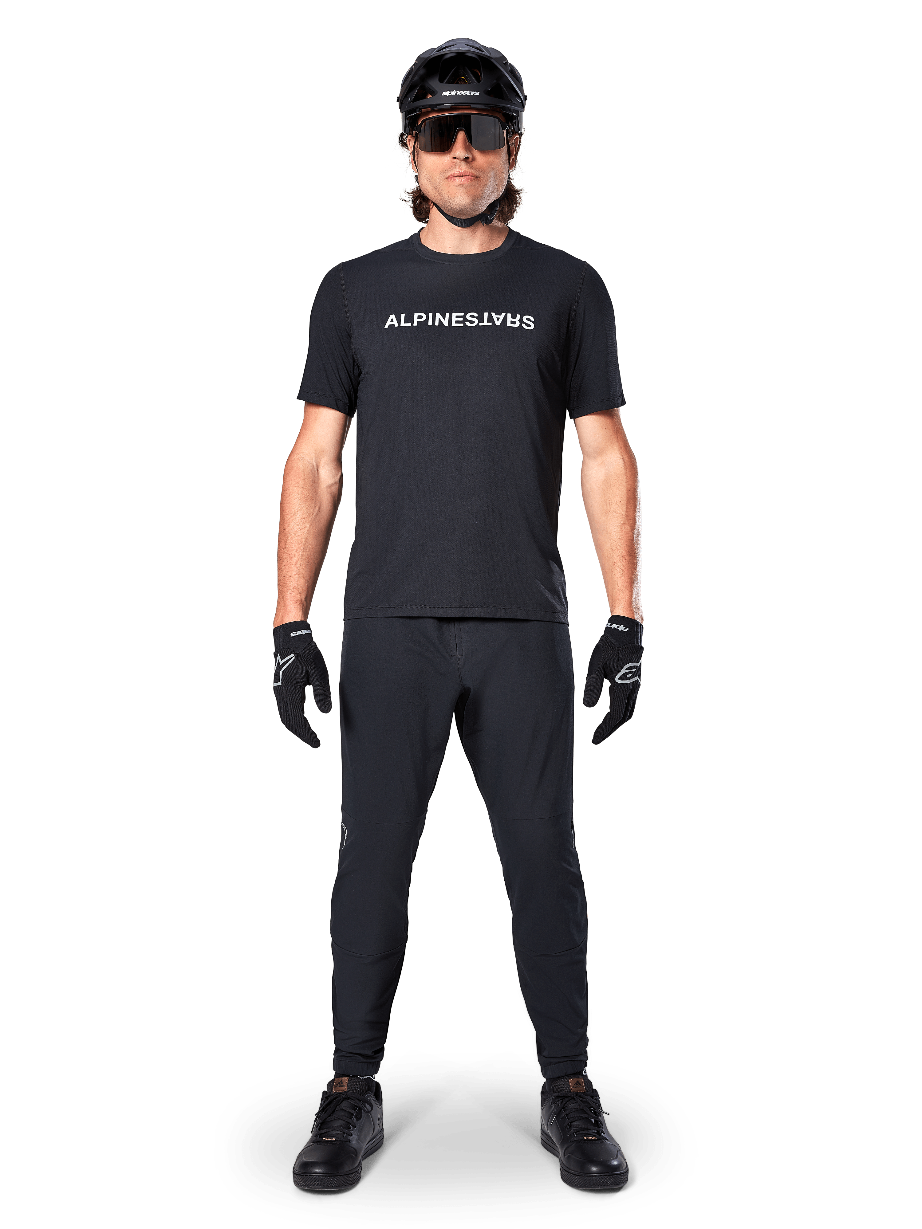 Ciclista di mountain bike che indossa la maglia Alpinestars A-Dura Switch a maniche corte in nero, con logo a scritta bianco sul petto, abbinata a pantaloni da MTB, casco e guanti neri coordinati, completo da ciclismo pronto per il trail.