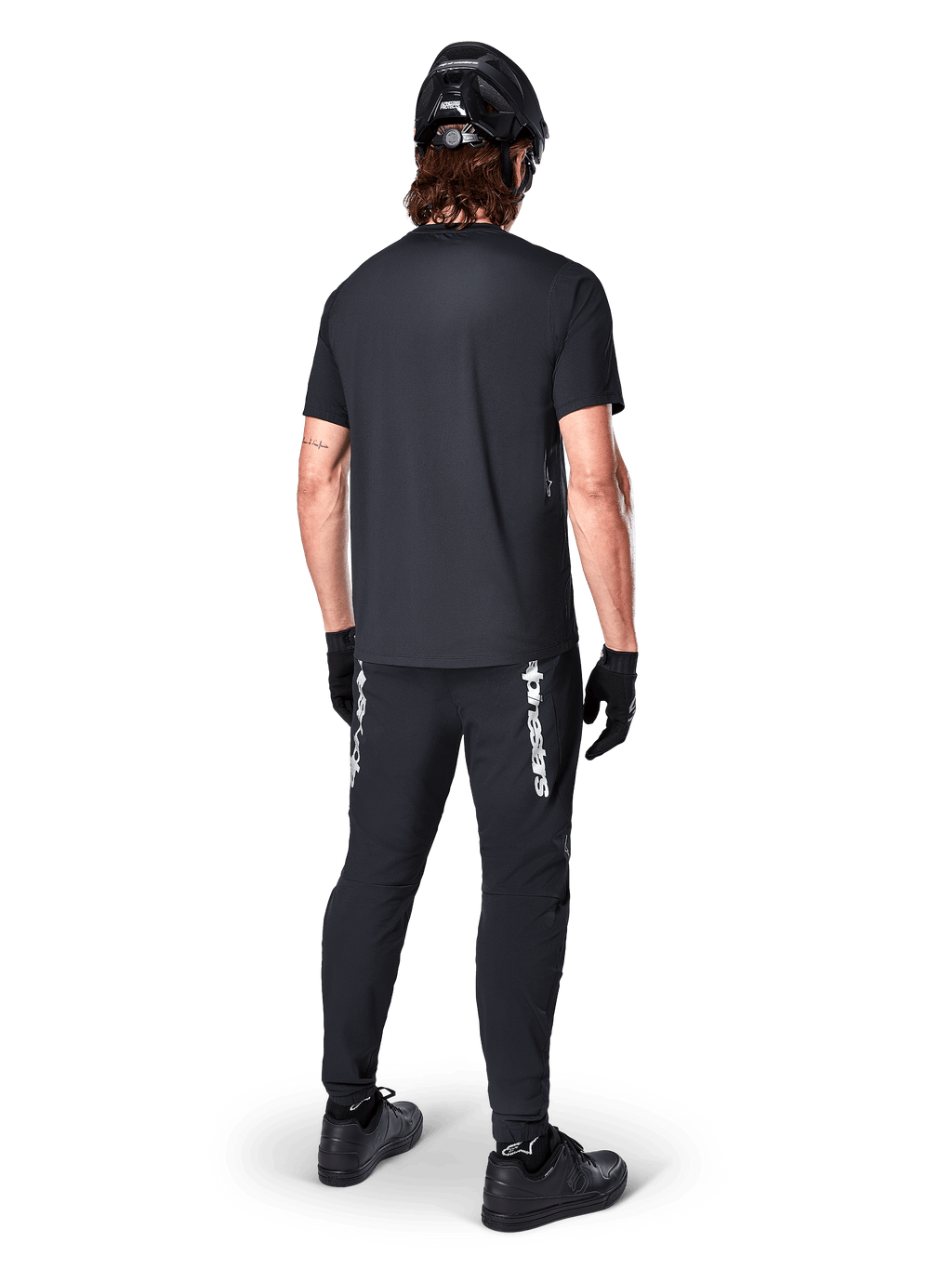 Alpinestars Maglia A-Dura Switch - Maniche Corte, Nero, vista posteriore di un ciclista con abbigliamento completo da mountain bike, inclusi pantaloni neri coordinati con logo bianco, casco nero e guanti, abbigliamento tecnico da ciclismo per trail riding