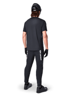 Alpinestars Maglia A-Dura Switch - Maniche Corte, Nero, vista posteriore di un ciclista con abbigliamento completo da mountain bike, inclusi pantaloni neri coordinati con logo bianco, casco nero e guanti, abbigliamento tecnico da ciclismo per trail riding