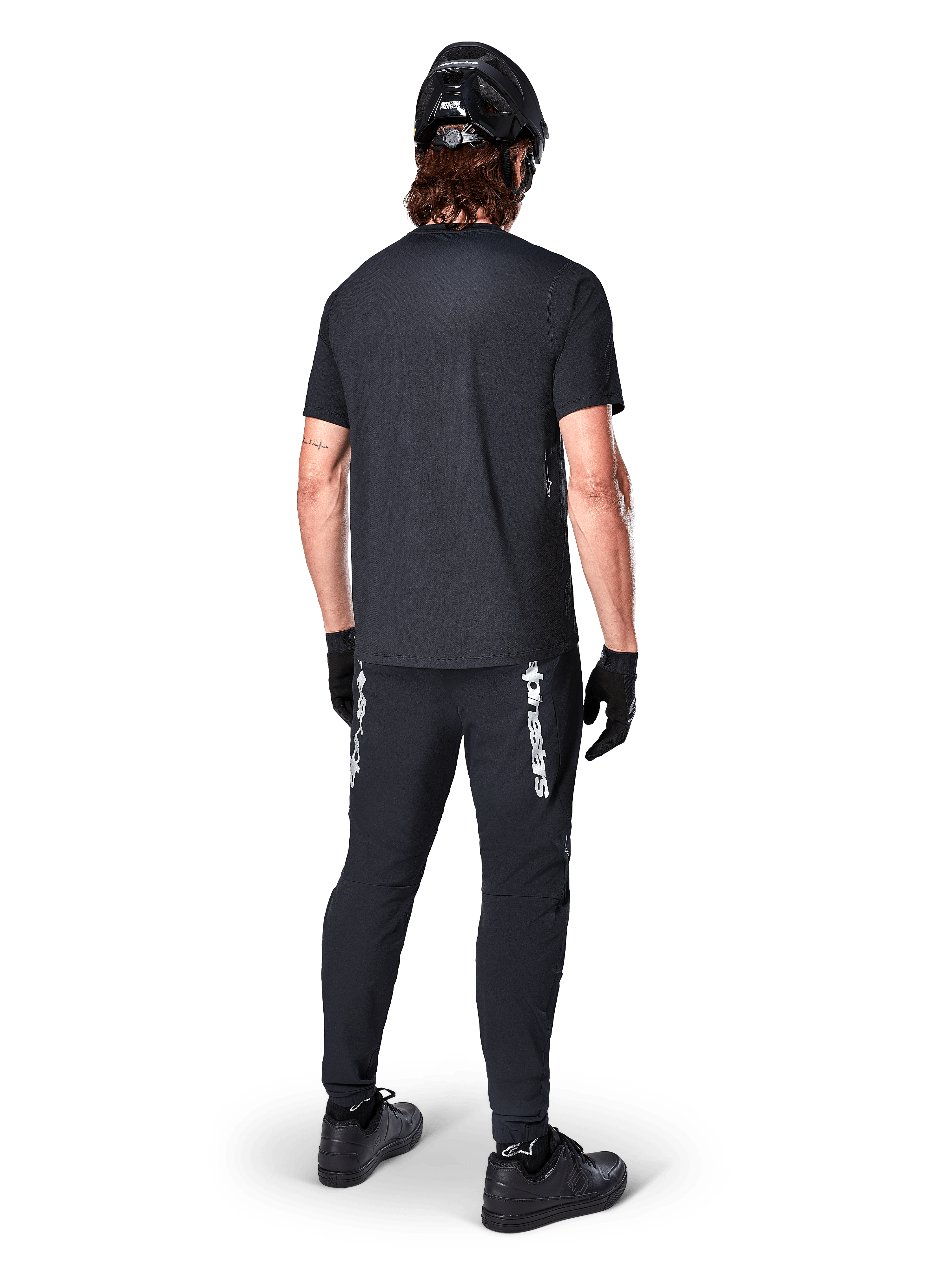 Alpinestars Maglia A-Dura Switch - Maniche Corte, Nero, vista posteriore di un ciclista con abbigliamento completo da mountain bike, inclusi pantaloni neri coordinati con logo bianco, casco nero e guanti, abbigliamento tecnico da ciclismo per trail riding