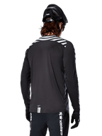 Maglia a maniche lunghe Alpinestars A-Dura Dri Oscar, Nero Riflettente, vista posteriore con strisce diagonali riflettenti sulle spalle e una coda più lunga, indossata con casco e guanti da MTB neri, equipaggiamento tecnico per mountain bike