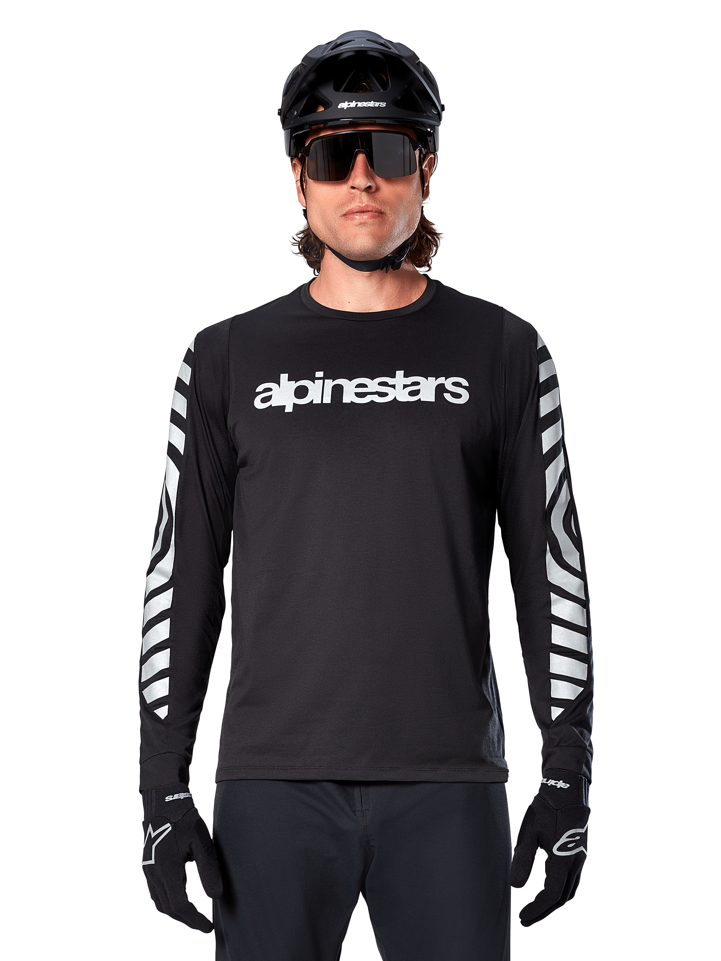 Ciclista uomo che indossa la maglia a maniche lunghe Alpinestars A-Dura Dri Oscar, Black Reflective, con logo Alpinestars bianco sul petto e grafiche riflettenti argento sulle maniche, abbinata a casco da MTB nero, occhiali da sole e guanti neri, abbigliamento tecnico da ciclismo