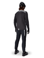 Ciclista di mountain bike con maglia a maniche lunghe Alpinestars A-Dura Dri Oscar in Black Reflective, vista da dietro, abbinata a pantaloni tecnici neri, casco e guanti, con grafiche a strisce riflettenti sulle spalle e coda posteriore allungata per il ciclismo fuoristrada.