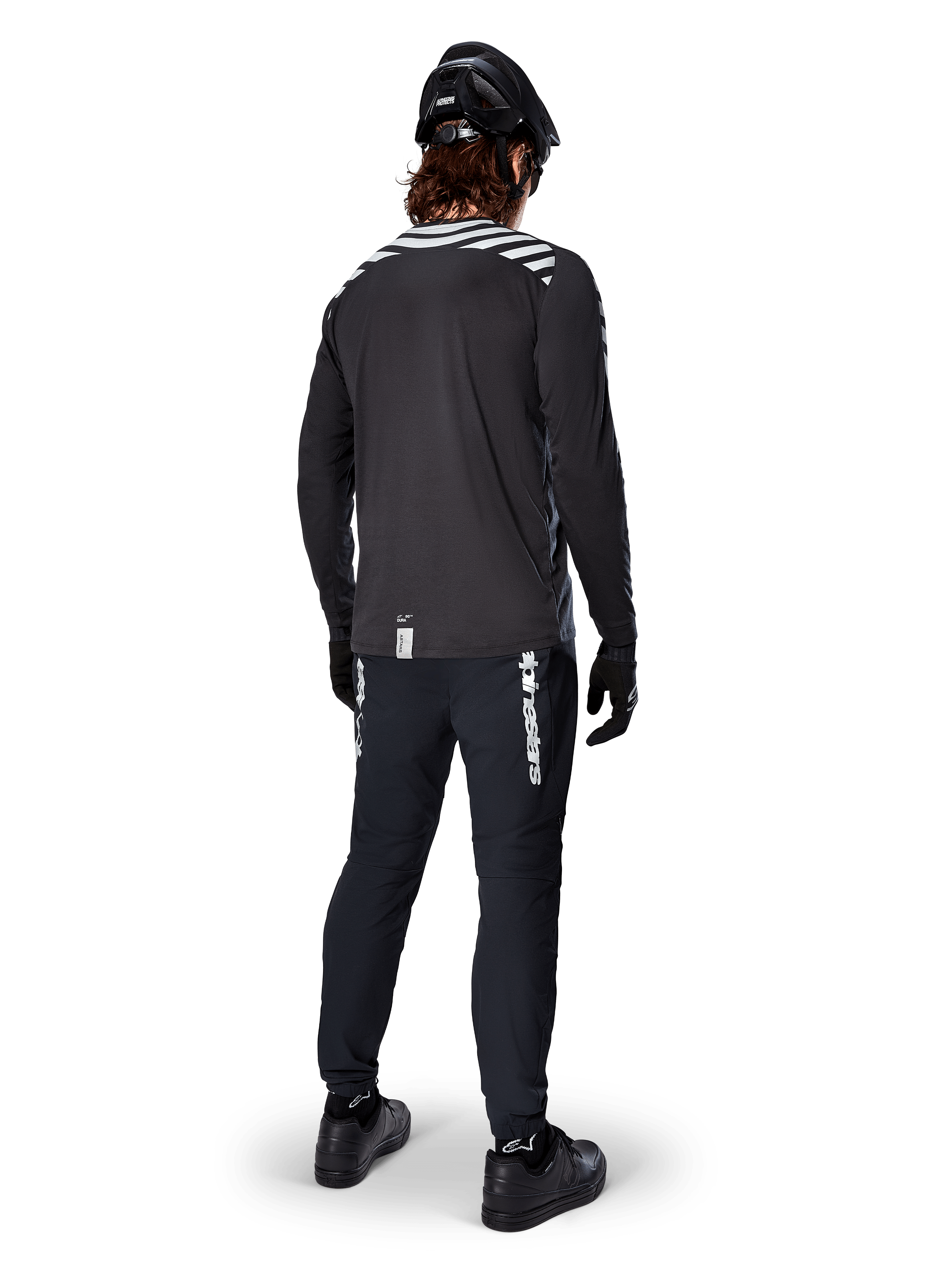 Ciclista di mountain bike con maglia a maniche lunghe Alpinestars A-Dura Dri Oscar in Black Reflective, vista da dietro, abbinata a pantaloni tecnici neri, casco e guanti, con grafiche a strisce riflettenti sulle spalle e coda posteriore allungata per il ciclismo fuoristrada.