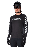 Alpinestars A-Dura Dri Oscar Jersey a maniche lunghe, maglia da ciclismo MTB, nero riflettente, con logo bianco sul petto e grafiche circolari riflettenti sulle maniche, indossata con casco nero e occhiali da sole, abbigliamento tecnico traspirante per mountain bike