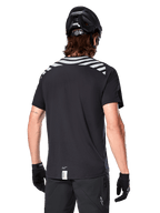 Maglia Alpinestars A-Dura Dri Oscar, maglia da MTB a maniche corte, Nero Riflettente, vista posteriore su modello con casco e guanti da ciclismo, con strisce riflettenti grigie sulle spalle e coda più lunga, tessuto tecnico traspirante per mountain bike