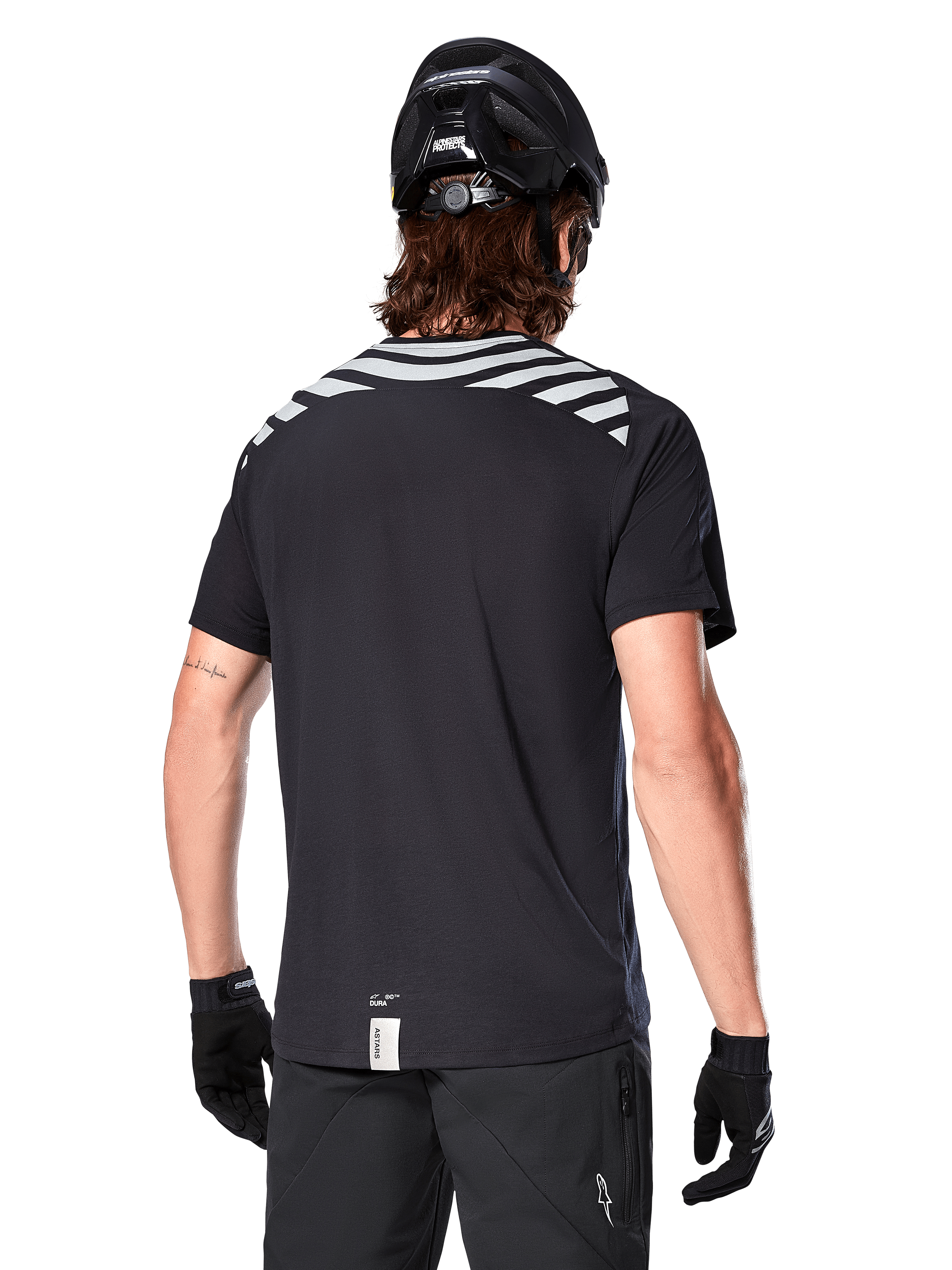 Maglia Alpinestars A-Dura Dri Oscar, maglia da MTB a maniche corte, Nero Riflettente, vista posteriore su modello con casco e guanti da ciclismo, con strisce riflettenti grigie sulle spalle e coda più lunga, tessuto tecnico traspirante per mountain bike