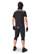 Maglia Alpinestars A-Dura Dri Oscar a maniche corte, nero riflettente, vista posteriore di un rider che indossa la maglia con pantaloncini da mountain bike neri, casco e ginocchiere, con grafiche riflettenti sulle spalle e design a coda lunga per MTB.