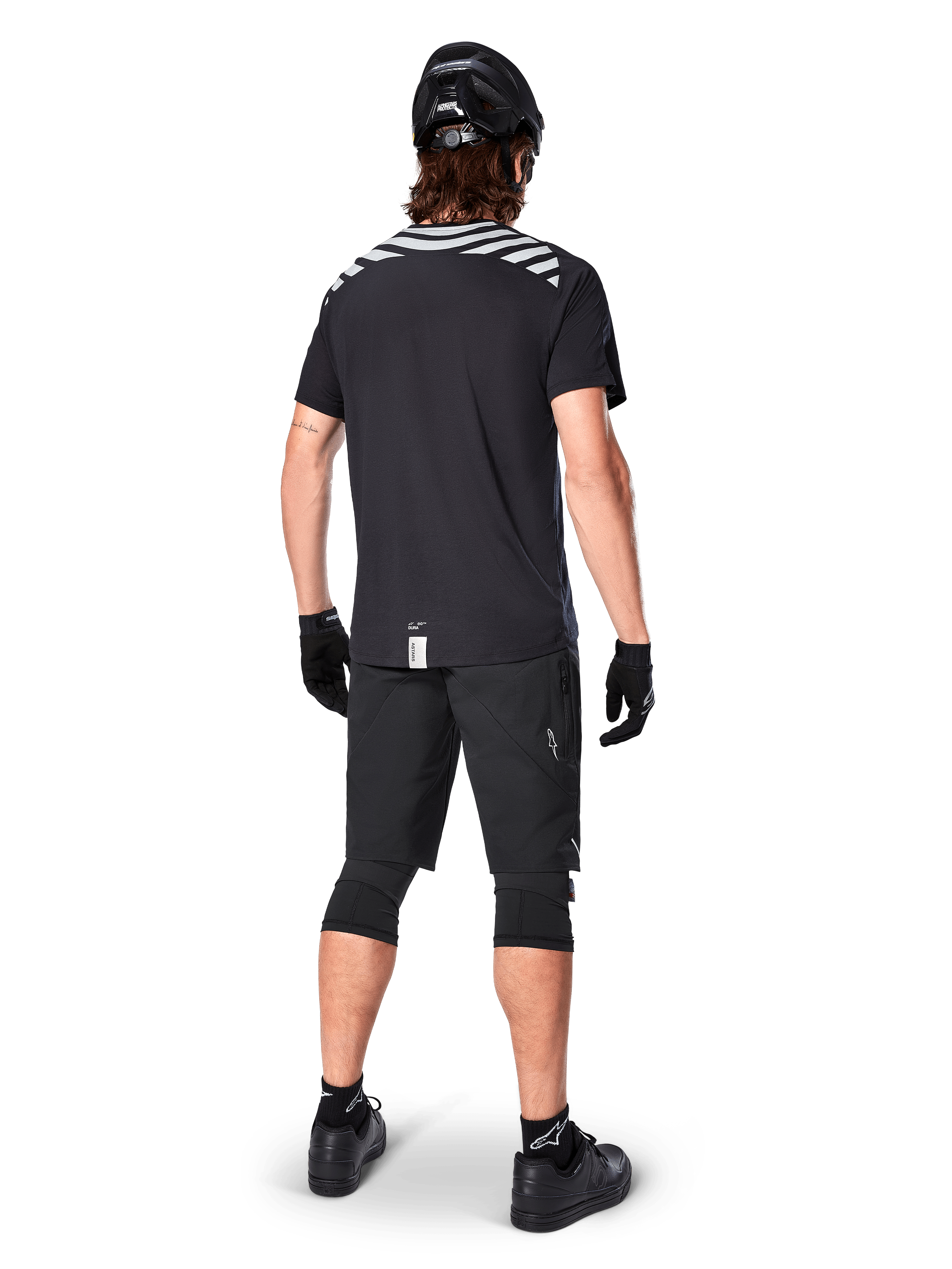 Maglia Alpinestars A-Dura Dri Oscar a maniche corte, nero riflettente, vista posteriore di un rider che indossa la maglia con pantaloncini da mountain bike neri, casco e ginocchiere, con grafiche riflettenti sulle spalle e design a coda lunga per MTB.