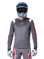 Ciclista MTB con maglia a maniche lunghe Alpinestars A-Supra Astar, grigio scuro con grafiche rosse sulle maniche e dettagli bianchi sulle spalle, pantaloni grigi coordinati con loghi a stella rossi, casco integrale blu e maschera, equipaggiamento da ciclismo fuoristrada coordinato