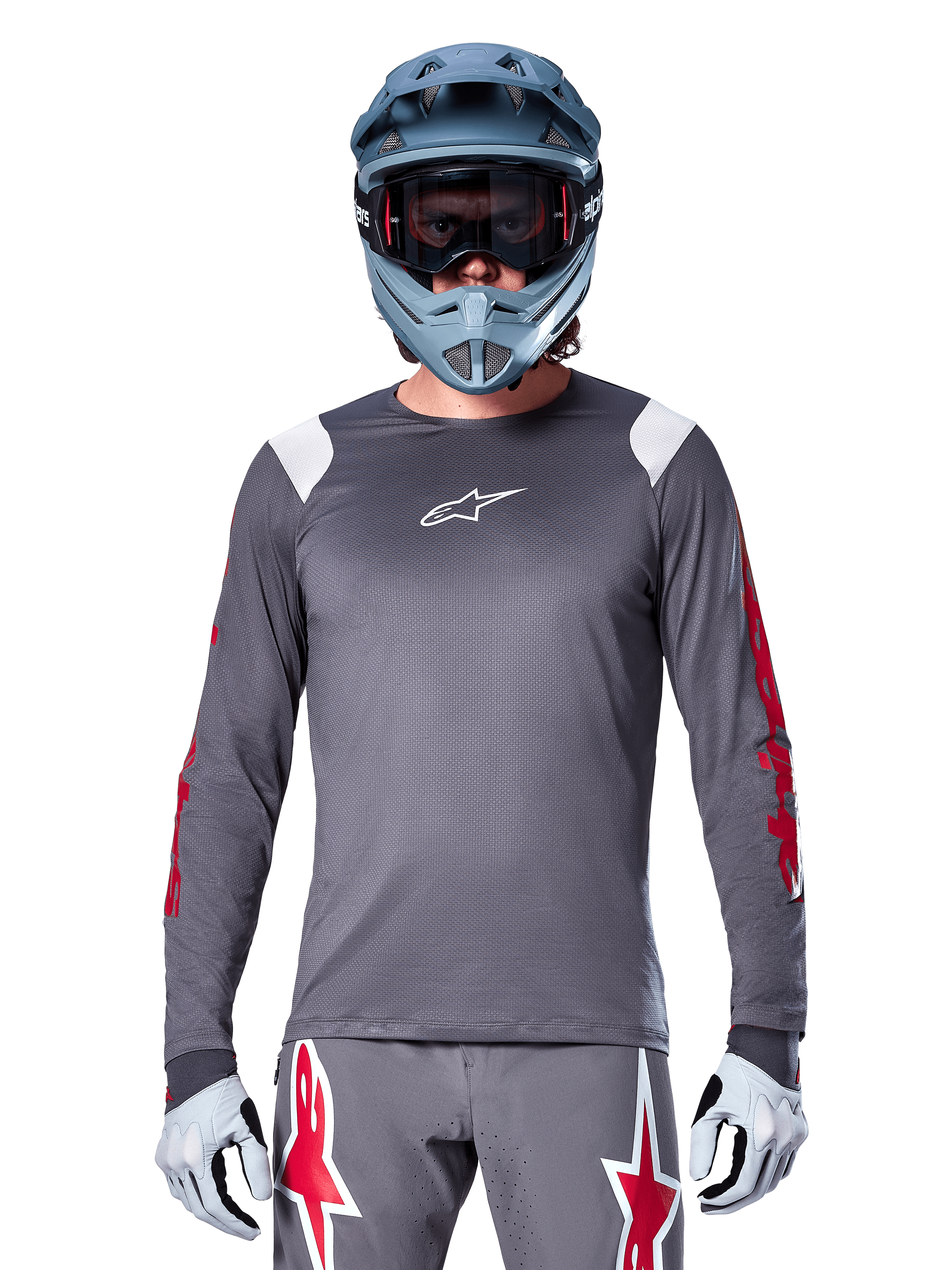 Ciclista MTB con maglia a maniche lunghe Alpinestars A-Supra Astar, grigio scuro con grafiche rosse sulle maniche e dettagli bianchi sulle spalle, pantaloni grigi coordinati con loghi a stella rossi, casco integrale blu e maschera, equipaggiamento da ciclismo fuoristrada coordinato