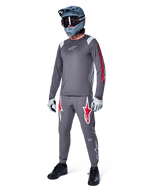 Maglia a maniche lunghe Alpinestars A-Supra Astar in grigio scuro, con branding rosso sulle maniche e logo bianco sul petto, indossata da un rider con pantaloni grigi coordinati, casco integrale grigio-blu e guanti tecnici, attrezzatura coordinata da mountain bike per il gravity.