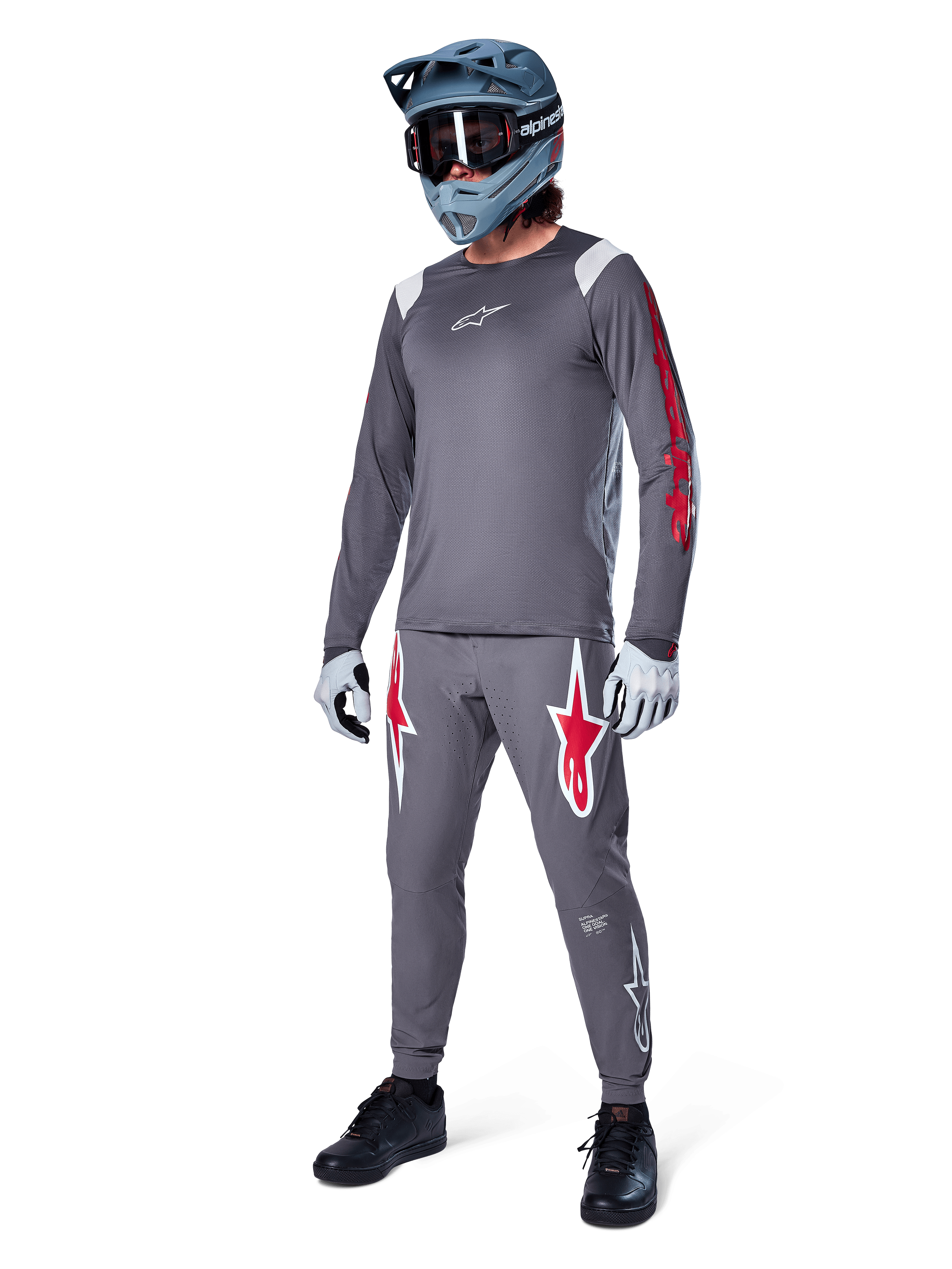 Maglia a maniche lunghe Alpinestars A-Supra Astar in grigio scuro, con branding rosso sulle maniche e logo bianco sul petto, indossata da un rider con pantaloni grigi coordinati, casco integrale grigio-blu e guanti tecnici, attrezzatura coordinata da mountain bike per il gravity.