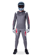 Ciclista MTB con maglia a maniche lunghe Alpinestars A-Supra Astar, grigio scuro con accenti rossi e logo bianco sul petto, pantaloni grigi coordinati con grafiche a stella rosse, casco integrale blu con maschera, guanti grigio chiaro e scarpe nere, completo tecnico professionale da mountain bike