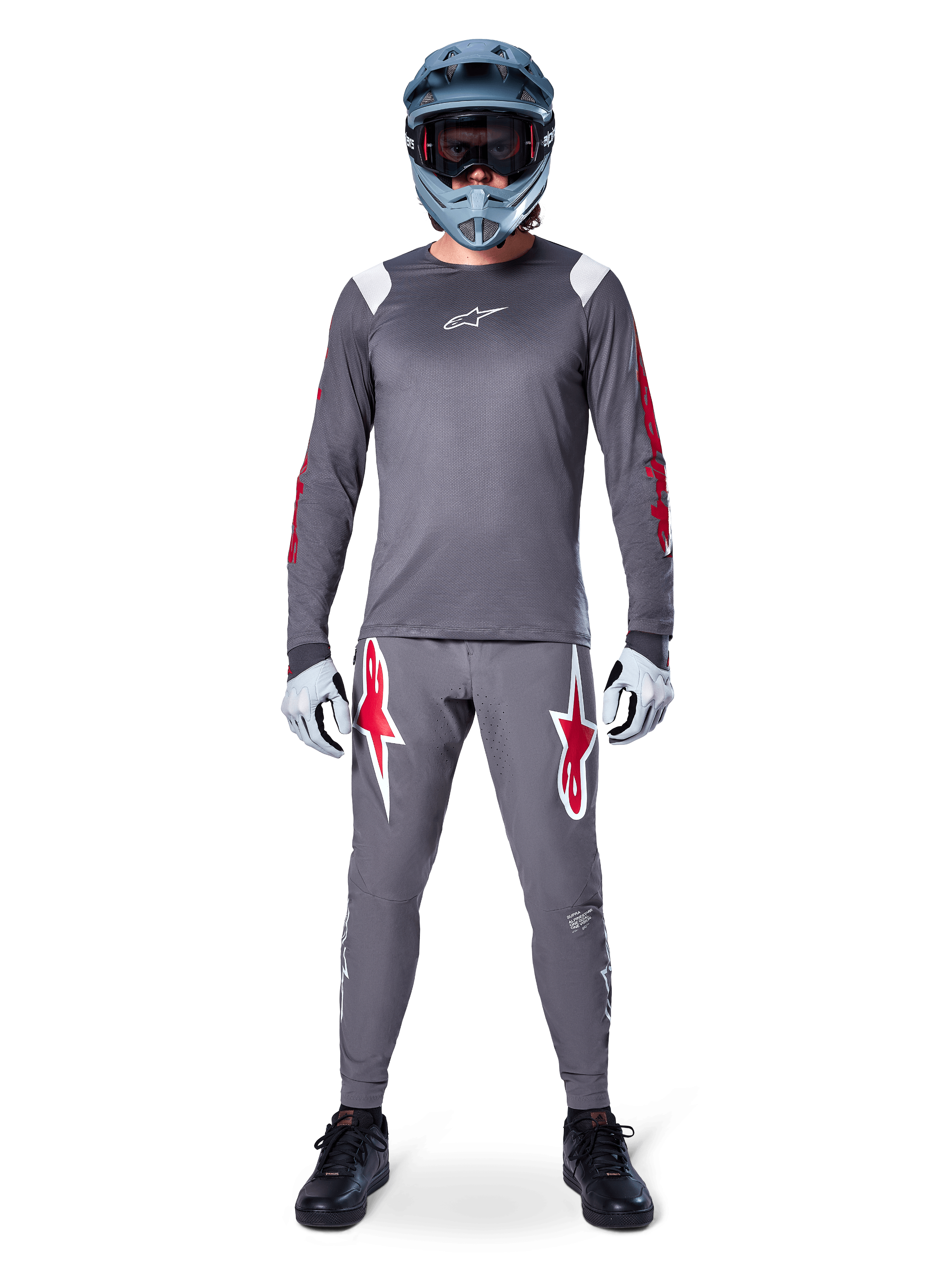 Ciclista MTB con maglia a maniche lunghe Alpinestars A-Supra Astar, grigio scuro con accenti rossi e logo bianco sul petto, pantaloni grigi coordinati con grafiche a stella rosse, casco integrale blu con maschera, guanti grigio chiaro e scarpe nere, completo tecnico professionale da mountain bike
