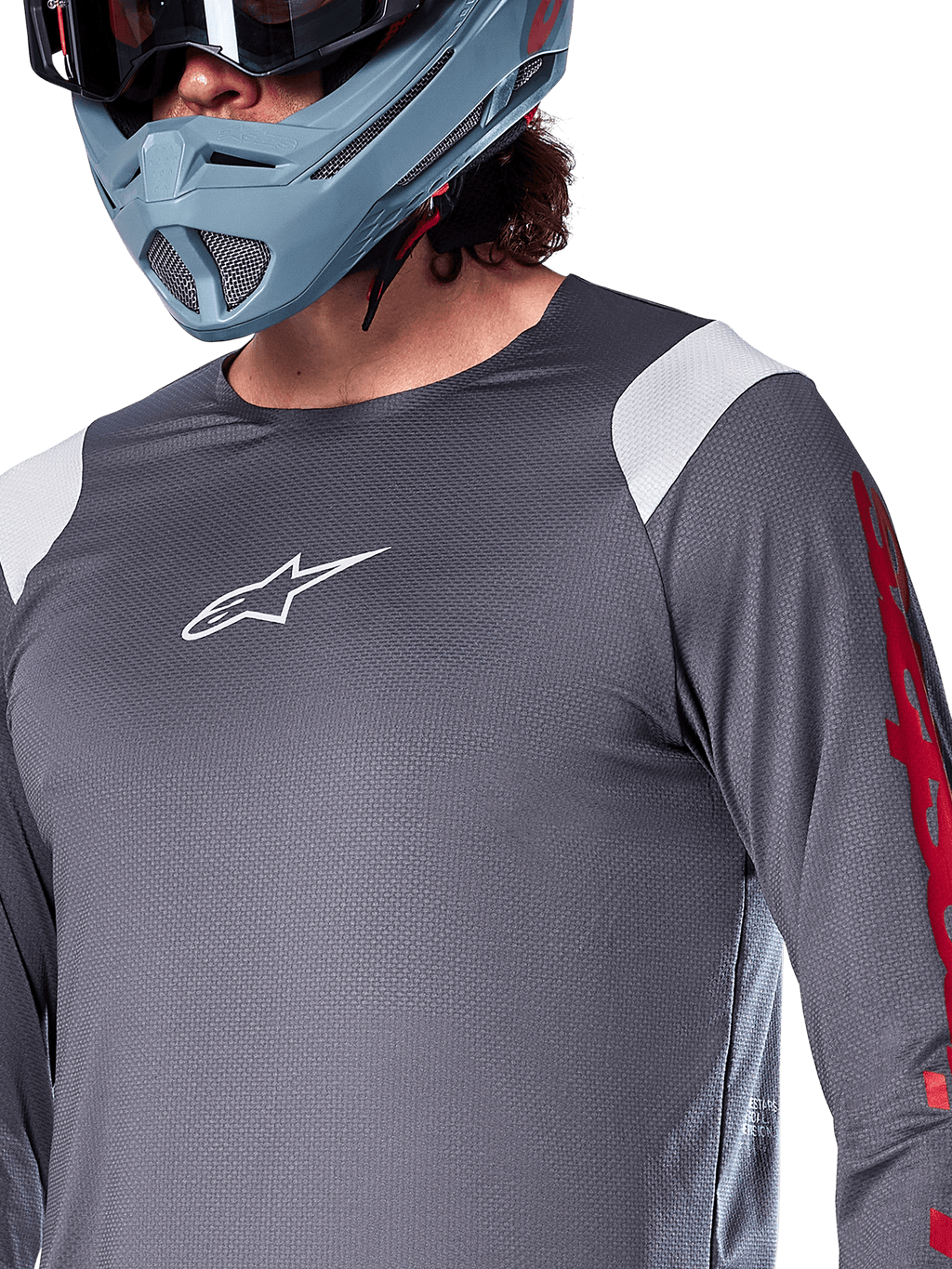 Alpinestars A-Supra Astar Jersey a maniche lunghe, grigio scuro con inserti bianchi sulle spalle e grafiche rosse sulle maniche, indossata con un casco integrale grigio-azzurro chiaro, abbigliamento tecnico per mountain bike