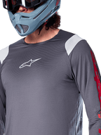 Alpinestars A-Supra Astar Jersey a maniche lunghe, grigio scuro con inserti bianchi sulle spalle e grafiche rosse sulle maniche, indossata con un casco integrale grigio-azzurro chiaro, abbigliamento tecnico per mountain bike