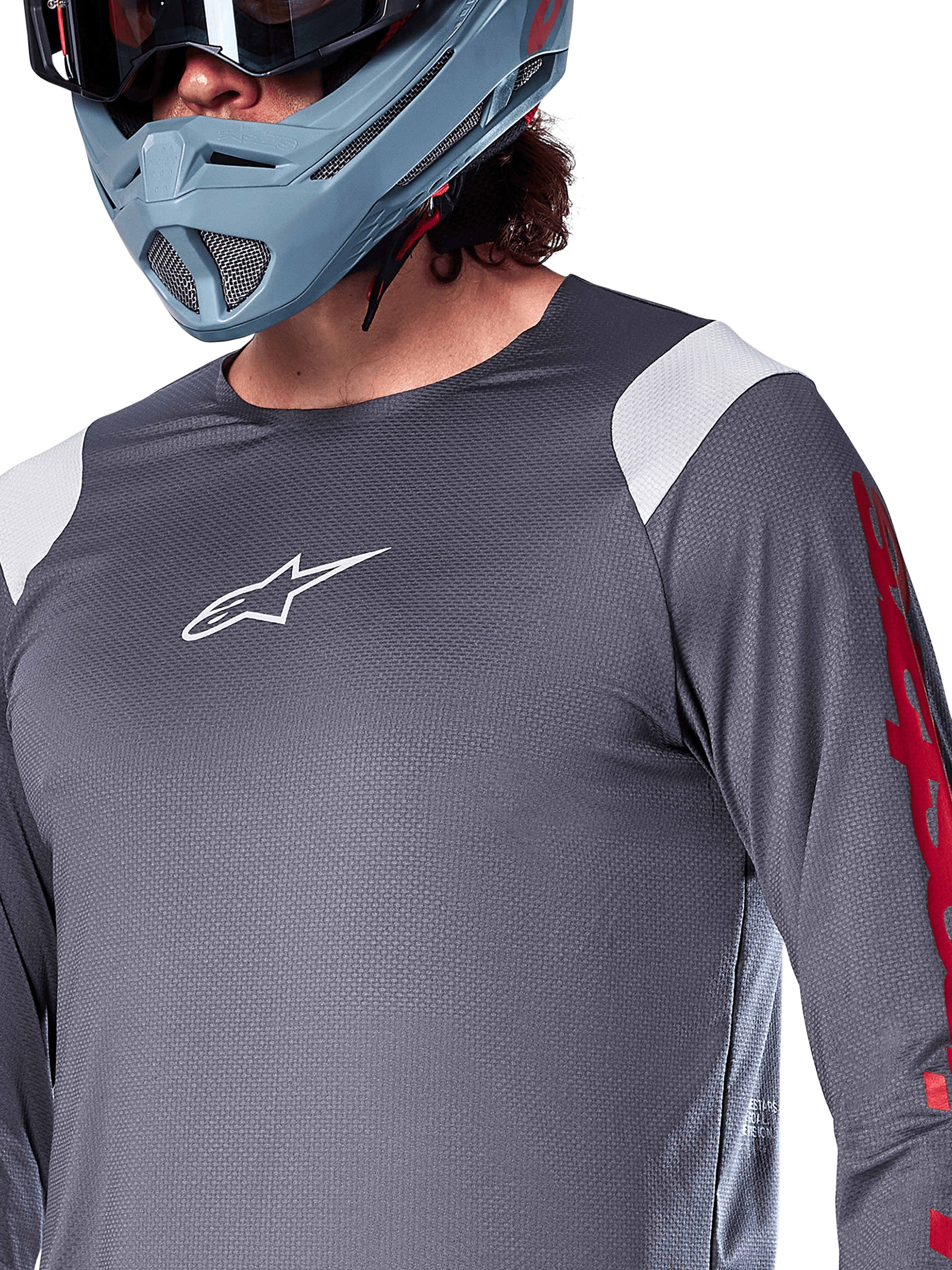 Alpinestars A-Supra Astar Jersey a maniche lunghe, grigio scuro con inserti bianchi sulle spalle e grafiche rosse sulle maniche, indossata con un casco integrale grigio-azzurro chiaro, abbigliamento tecnico per mountain bike