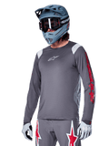 Alpinestars A-Supra Astar Jersey a maniche lunghe, maglia da mountain bike MTB, grigio scuro con accenti rossi e bianchi, vestibilità performance, indossata con pantaloni grigi coordinati con grafiche a stella rosse e bianche, completo da ciclismo off-road con casco integrale e guanti