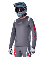 Alpinestars A-Supra Astar Jersey a maniche lunghe, maglia da mountain bike MTB, grigio scuro con accenti rossi e bianchi, vestibilità performance, indossata con pantaloni grigi coordinati con grafiche a stella rosse e bianche, completo da ciclismo off-road con casco integrale e guanti