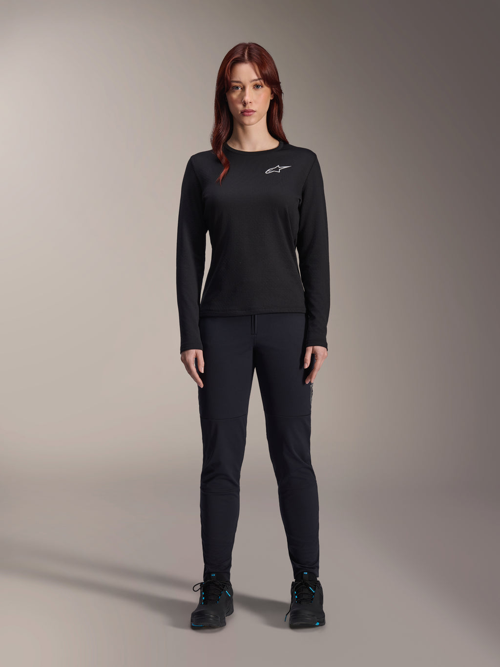 Alpinestars Stella A-Dura Thermal Jersey, maglia da mountain bike a maniche lunghe da donna in nero, con logo bianco sul petto, indossata con abbigliamento nero coordinato e scarpe da ciclismo, ritratto in studio.